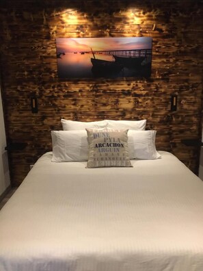 A bed with white bedding and a pillow with text, set against a wooden wall with a sunset painting above.