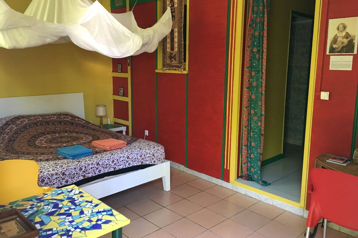 A room with a bed, a small table, a chair, a hanging canopy, and a door leading to another room.