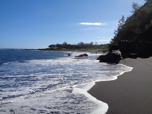 Una playa de arena oscura, olas y una zona boscosa al fondo.