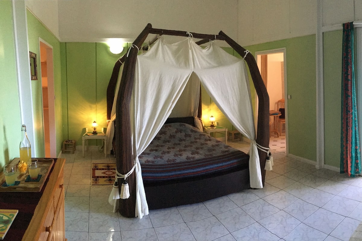 A room with a four-poster bed, a small table with a lamp, and a view of the outside through a sliding glass door.