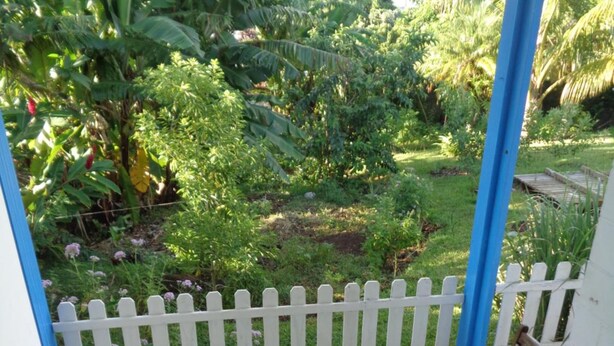 A wooden table and two chairs on a tiled patio with a white picket fence and lush greenery in the background. - L'Arbre de Vie (Saint-Joseph)
