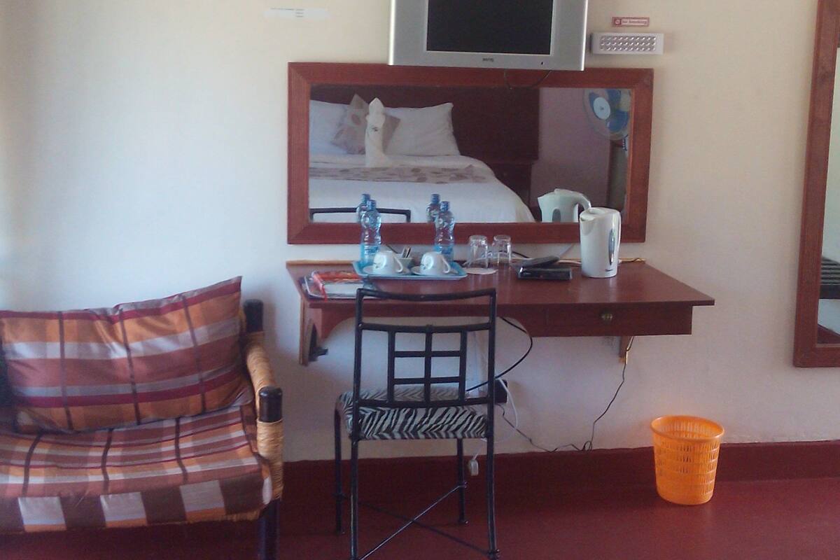 Una habitación de hotel con una cama, un escritorio con una computadora, una silla, una mesita con botellas de agua, un cesto de basura y una alfombra estampada.