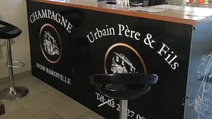 A bar area with a counter labeled "CHAMPAGNE Urbain Père & Fils" and a phone number. There are two stools and a wine bottle on the counter. - Champagne Urbain (Baroville)