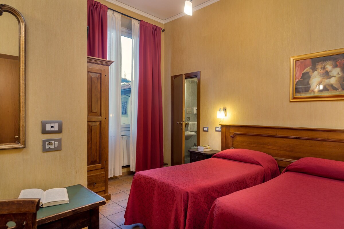A hotel room with two beds, a wooden headboard, a painting, a mirror, a desk with a book, and a window with curtains.
