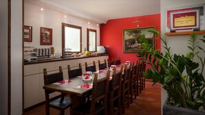 Un comedor con una larga mesa de madera dispuesta para una comida, rodeada de sillas. Se observa una zona de cocina con una encimera y varios objetos encima. Una planta grande es visible en primer plano.