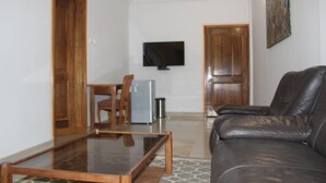 A living room with a leather sofa, a glass coffee table, a TV mounted on the wall, and a wooden door leading to another room. - Maison D Hote Villy (Ouagadougou)