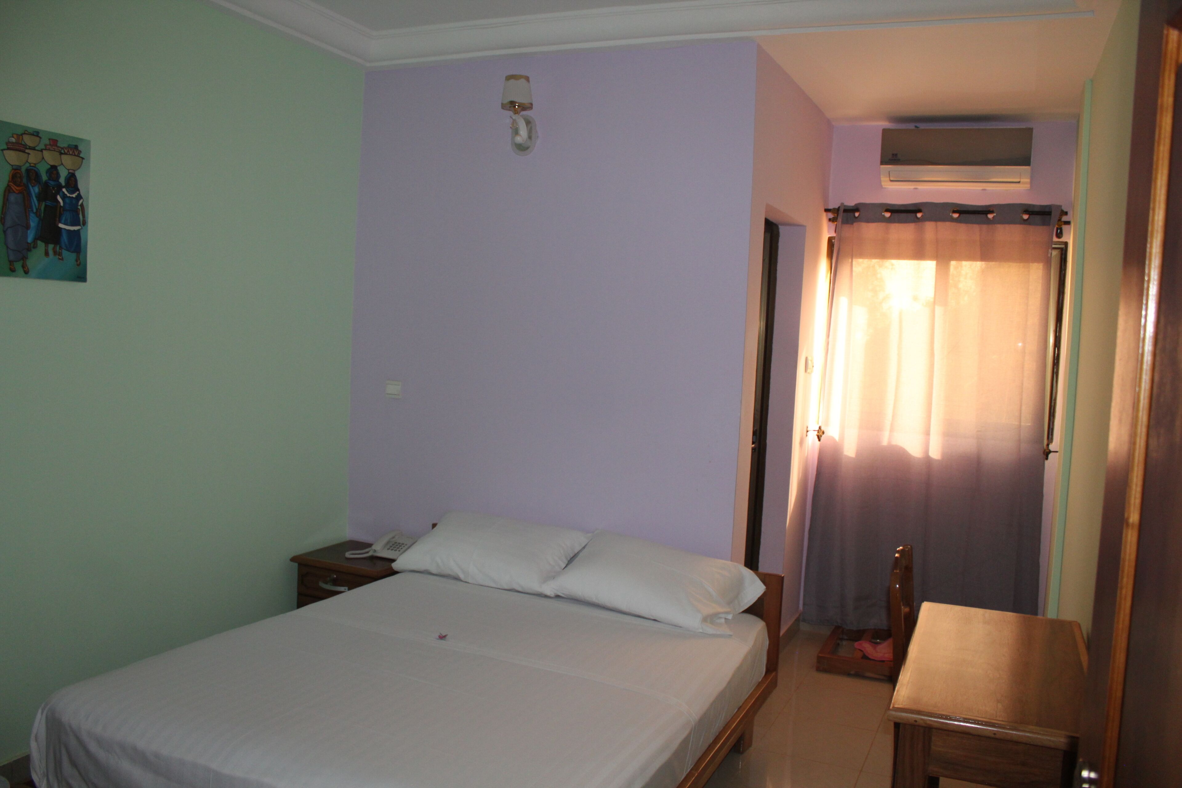 A bedroom with a bed, a bedside table, a nightstand, a wall-mounted lamp, and a painting on the wall.