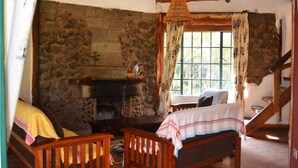 A rustic room with a stone wall, a fireplace, wooden furniture, and a window with curtains. - Oleitiko Cottages (Naivasha)