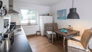 A compact kitchen with modern appliances, a dining area with wicker furniture, and a view of greenery outside. - Diekhuus Arngast, Apartment Butendiek - Apartment Butendiek in the Diekhuus (Varel)