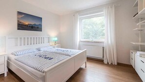 A bedroom with a bed, bedside tables, a window with curtains, a radiator, and a wall-mounted shelf. - Diekhuus Arngast, Apartment Butendiek - Apartment Butendiek in the Diekhuus (Varel)