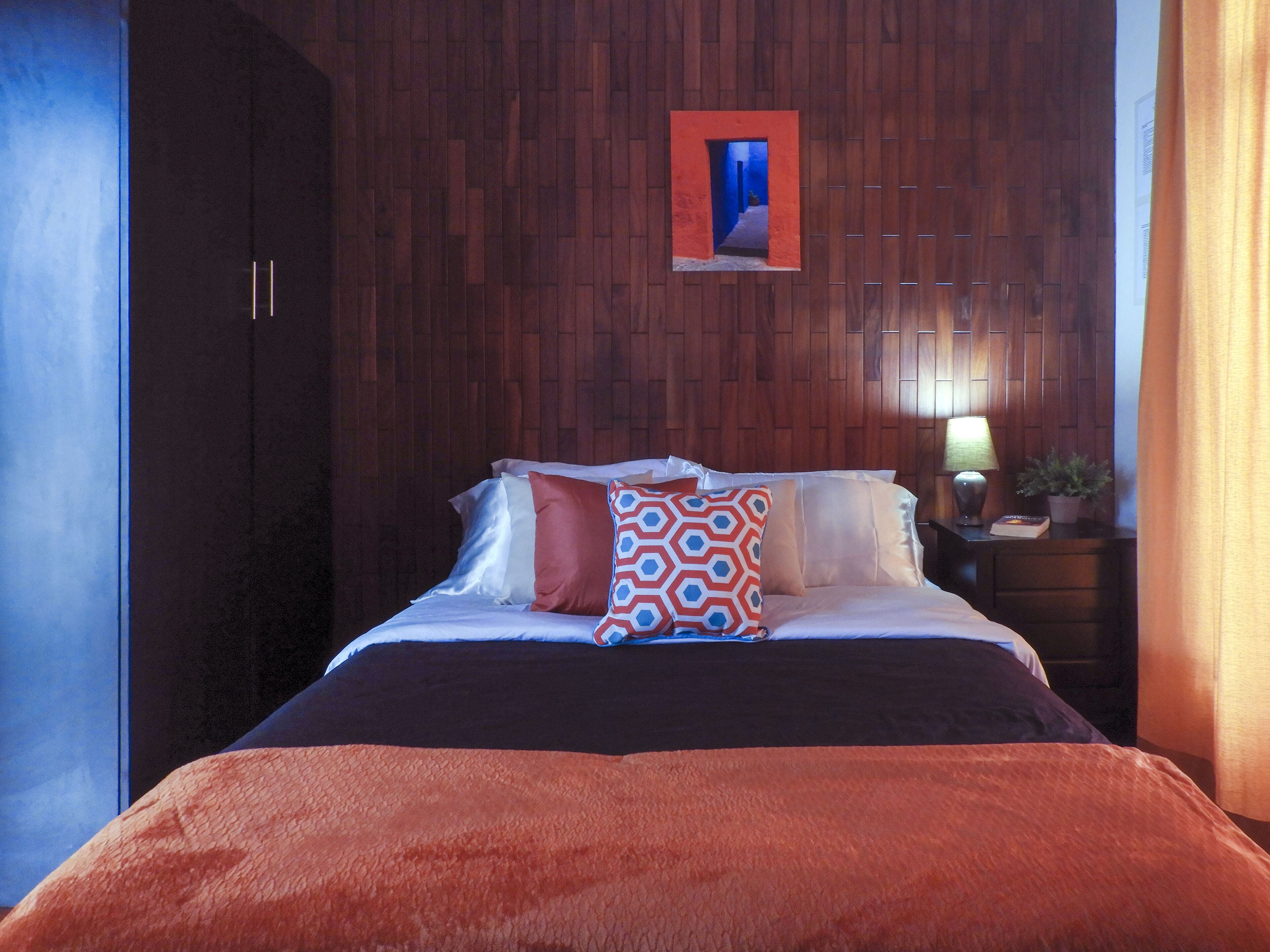 A hotel room with a wooden headboard, a bed with multiple pillows, a nightstand with a lamp and a plant, and a framed picture on the wall.