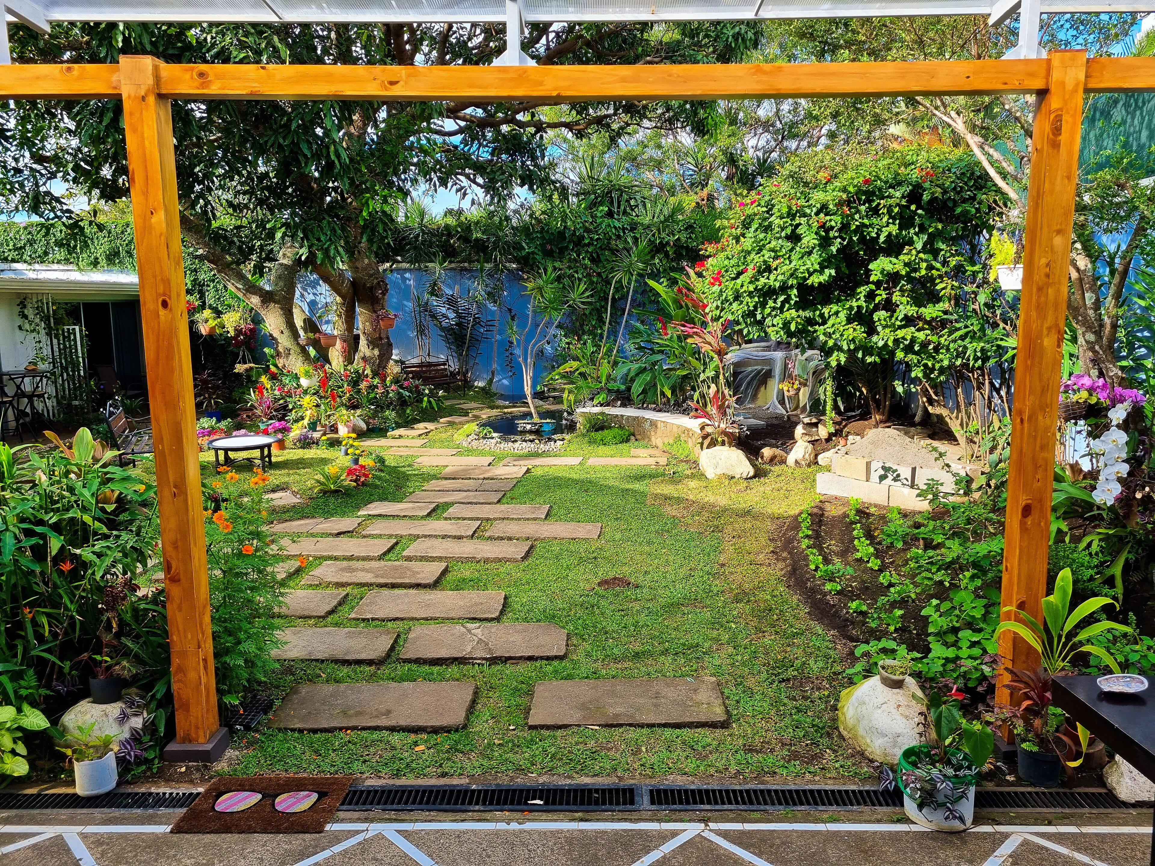 Un jardín con una pérgola de madera, piedras que forman un sendero y diversas plantas en macetas.