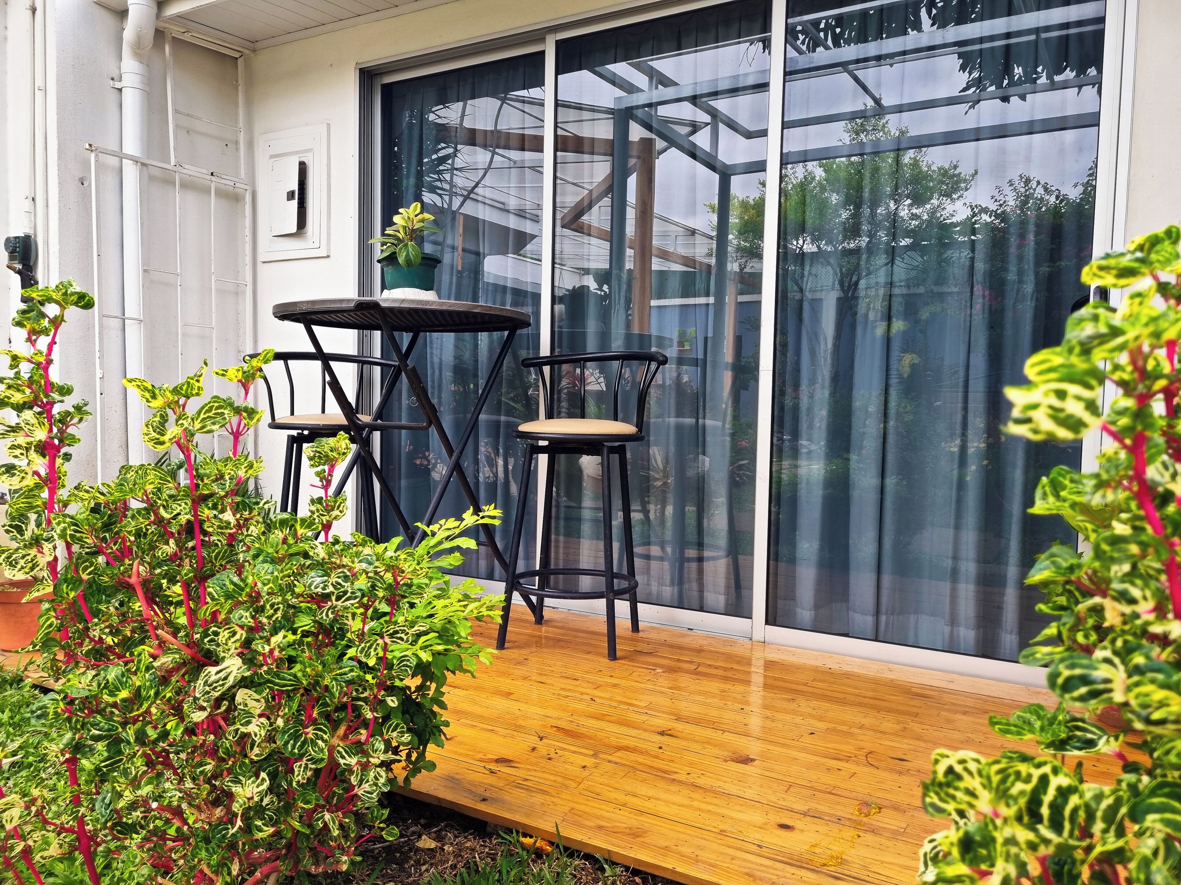 Una terraza de madera con dos sillas negras y una mesita, rodeada de macetas con plantas de hojas variegadas.