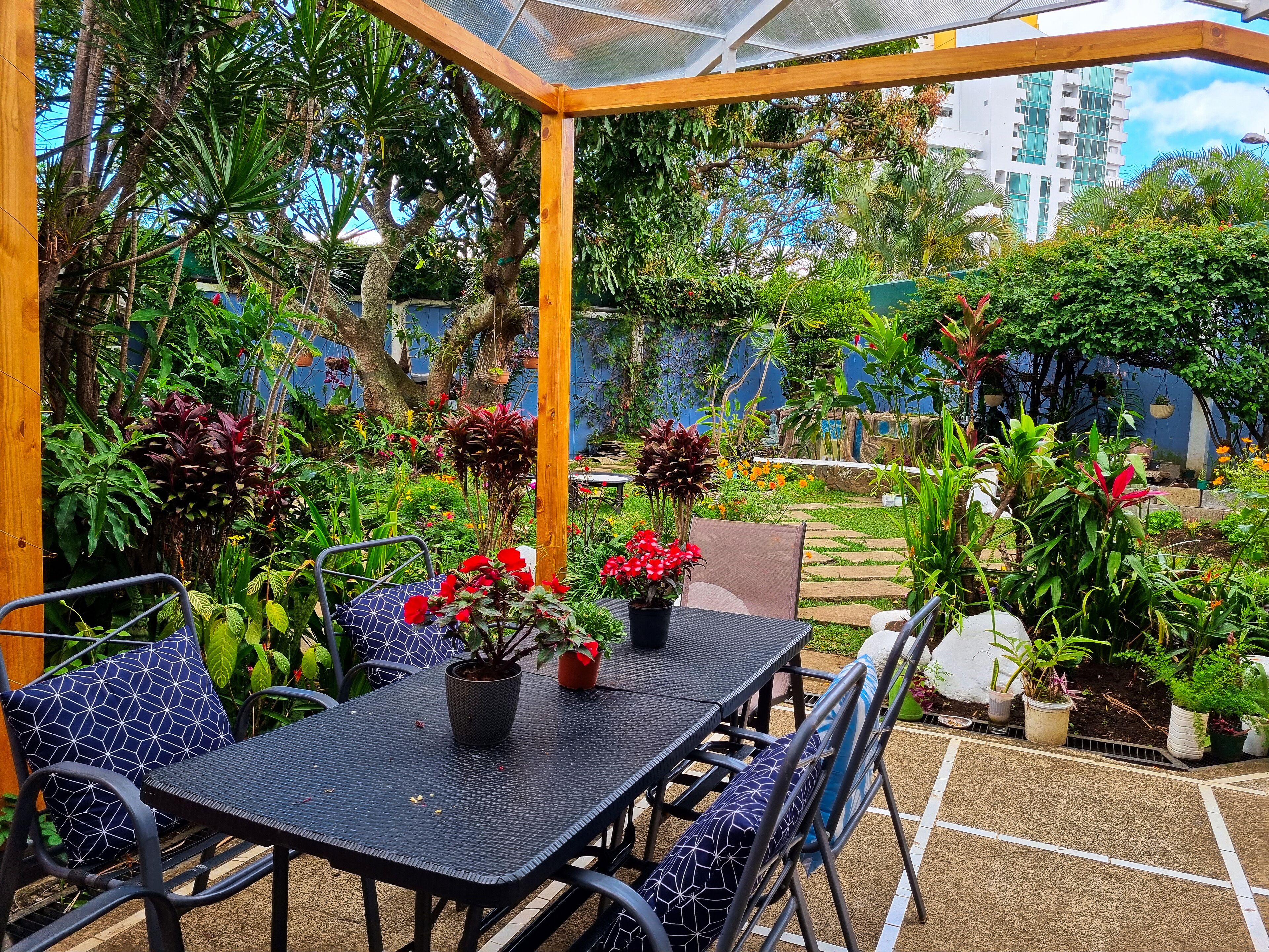 Un patio al aire libre con mesa y sillas, rodeado de abundante follaje y plantas en macetas.