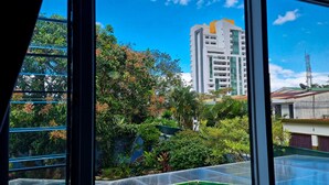 A view through a window of a building with a green roof, surrounded by lush greenery and a tall white building with a yellow accent in the background. - Casa Jardin Del Mango (San José)