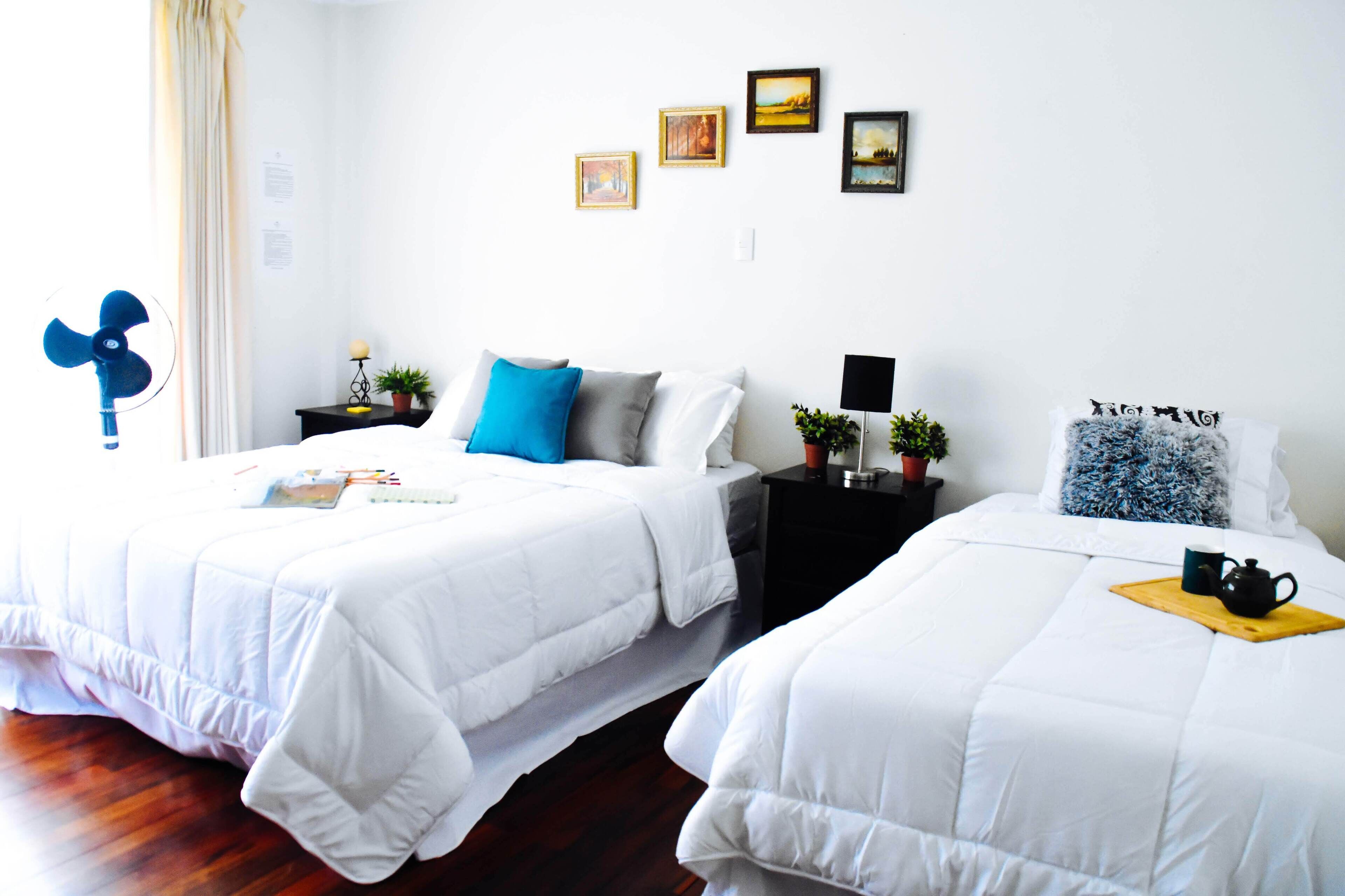 A hotel room with two beds, white bedding, a blue fan, and artwork on the wall.