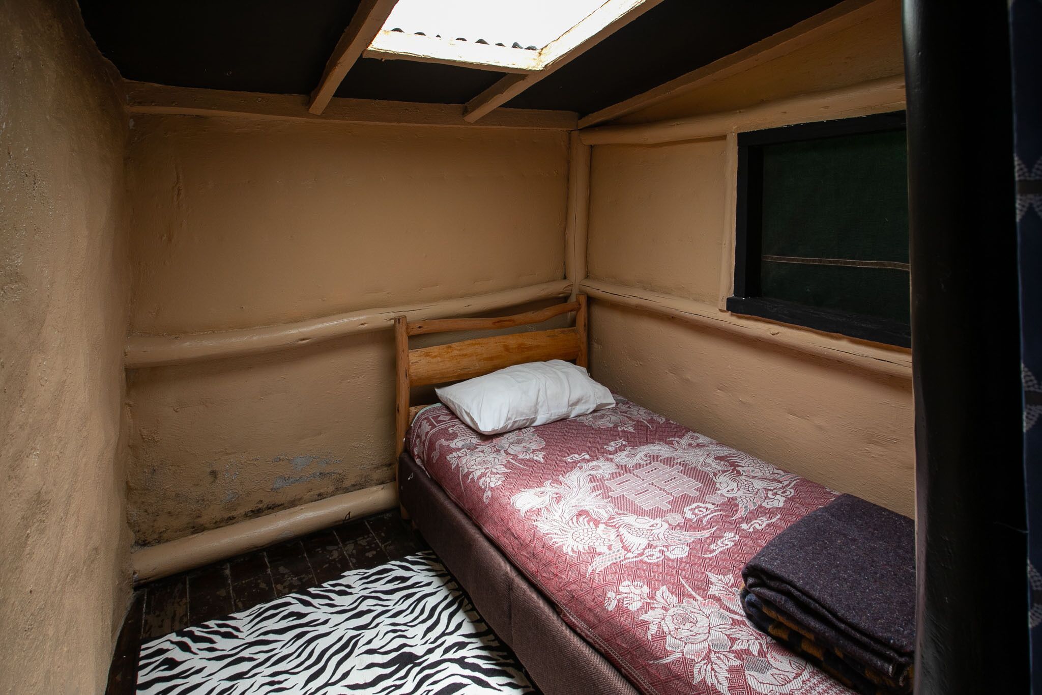 A small, single-bed room with a wooden headboard, a zebra-patterned rug, a red floral bedspread, and a window with a black frame.