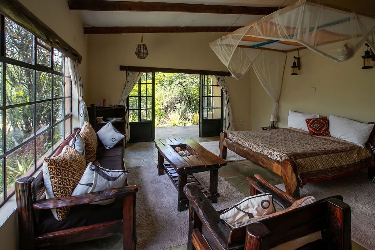 A room with a bed, a wooden bench, and a table. There is a window with curtains and a view of greenery outside.