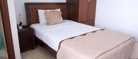 Una cama bien hecha con cabecera de madera, una mesita de noche con una planta y un armario con espejo.