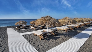 A beach with rows of sun loungers and thatched umbrellas, a wooden walkway, and the ocean in the background. - Meltemi Blu - Adults Only (Santorini)