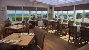 A sunlit dining area with wooden tables and chairs, large windows offering a view of the sea, and a ceiling fan. - Port Charlotte Hotel (Islay Island)