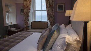 A hotel room with a bed, a chair, a desk, a lamp, and a window with curtains. - Port Charlotte Hotel (Islay Island)