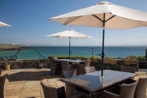 An outdoor patio with wicker furniture and large umbrellas, overlooking a coastal view with a lighthouse in the distance.
