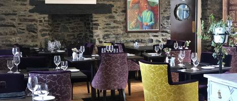 A restaurant with stone walls, wooden tables, purple and yellow chairs, and a painting of a horse.