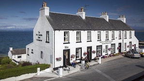 A white building with a black roof, named 'The Port Charlotte Hotel & Restaurant'. It has multiple windows, a bar area with outdoor seating, and is located near a body of water. - Port Charlotte Hotel (Islay Island)