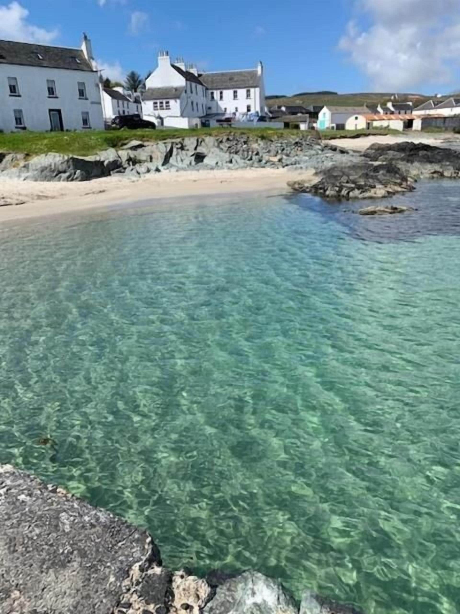 A coastal scene with clear water, a sandy beach, and white buildings in the background.