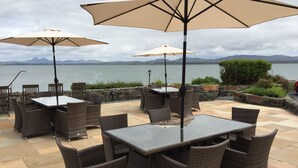 An outdoor patio with wicker furniture and large umbrellas, overlooking a body of water and mountains. - Port Charlotte Hotel (Islay Island)