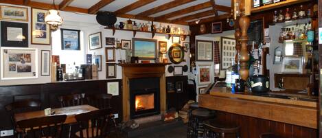 A pub with wooden beams, a fireplace, bar area with stools, and various framed pictures on the walls.
