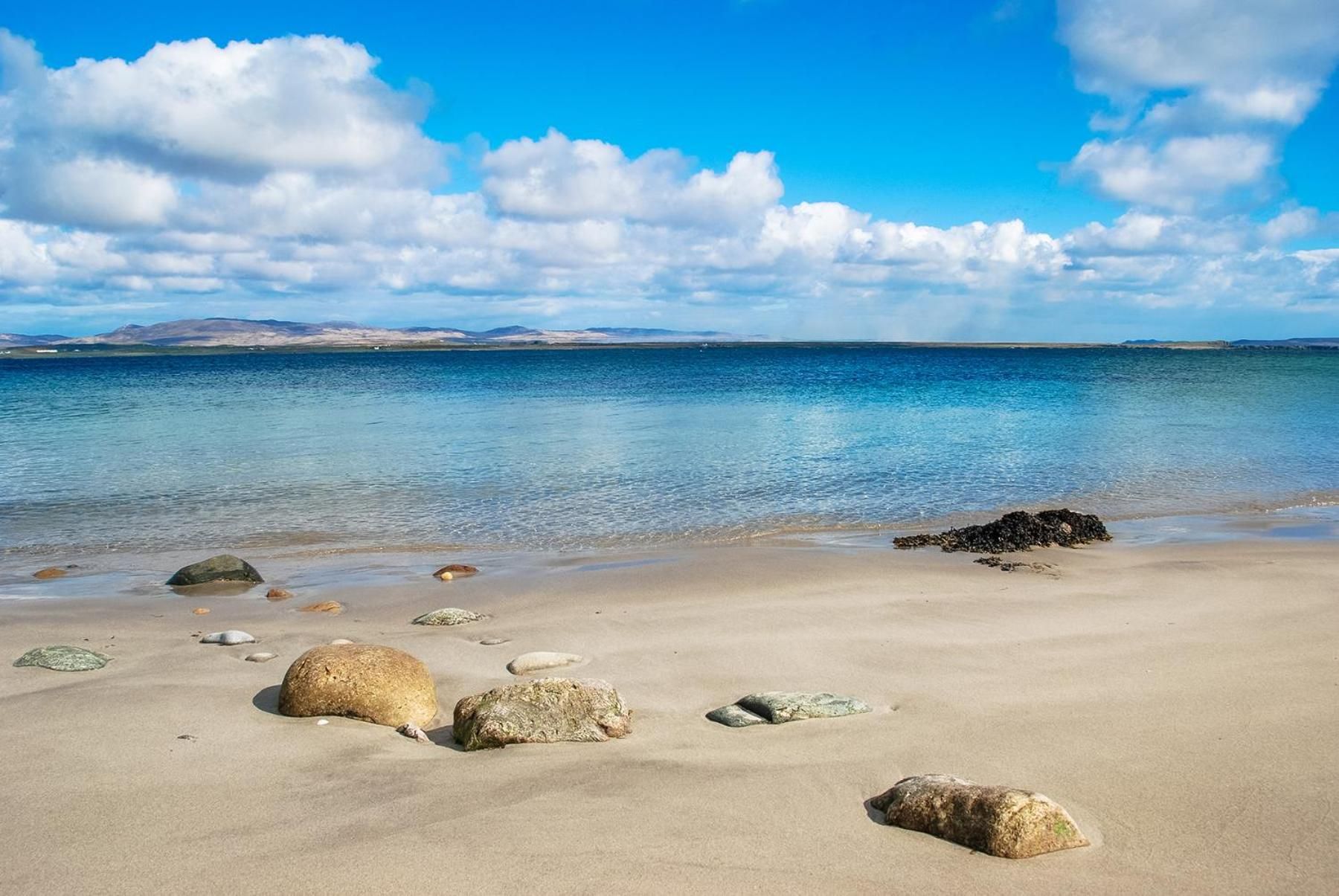 A beach with clear water, scattered rocks, and distant mountains under a blue sky.
