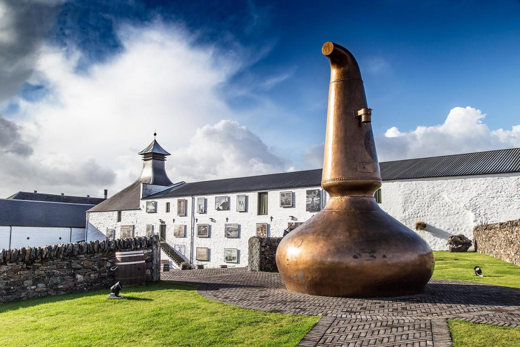 A large copper distillation still in front of a white building with a stone wall and a tower.