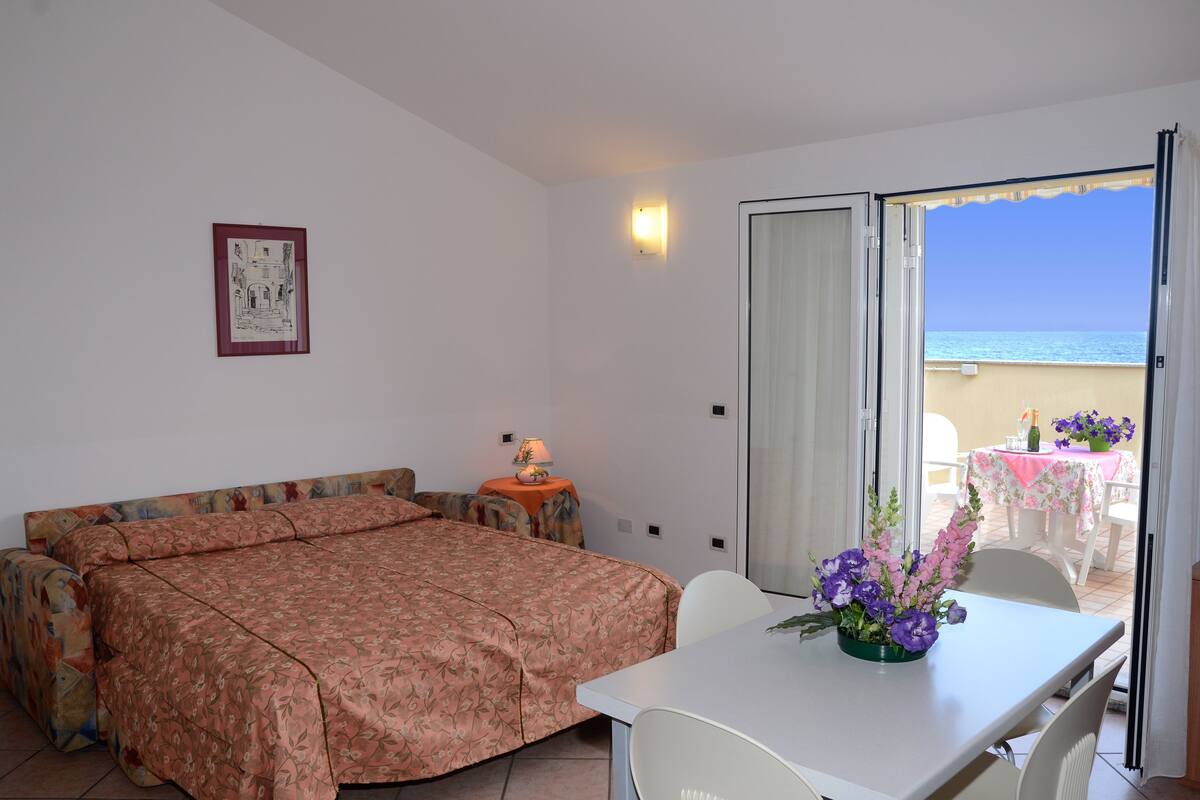 Habitación de hotel con una cama, una mesa con un jarrón de flores, una lámpara y vista al mar a través de un ventanal corredera.