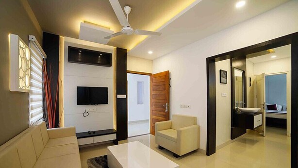 A modern living room with a sofa, a white coffee table, a TV mounted on the wall, a ceiling fan, and a bathroom visible through an open door. - Ambalath Maple Leaves - Hostel (Chavakkad)