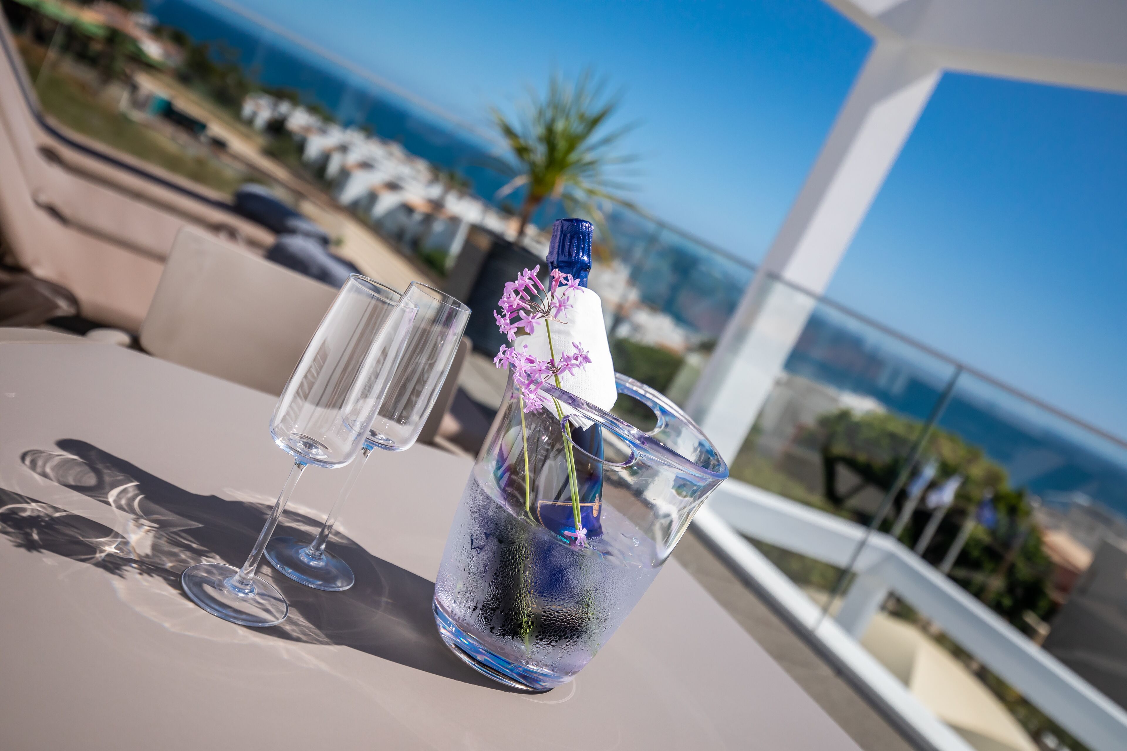 A table with a glass pitcher containing flowers and a clear liquid, two empty glasses, and a view of the ocean and buildings in the background.