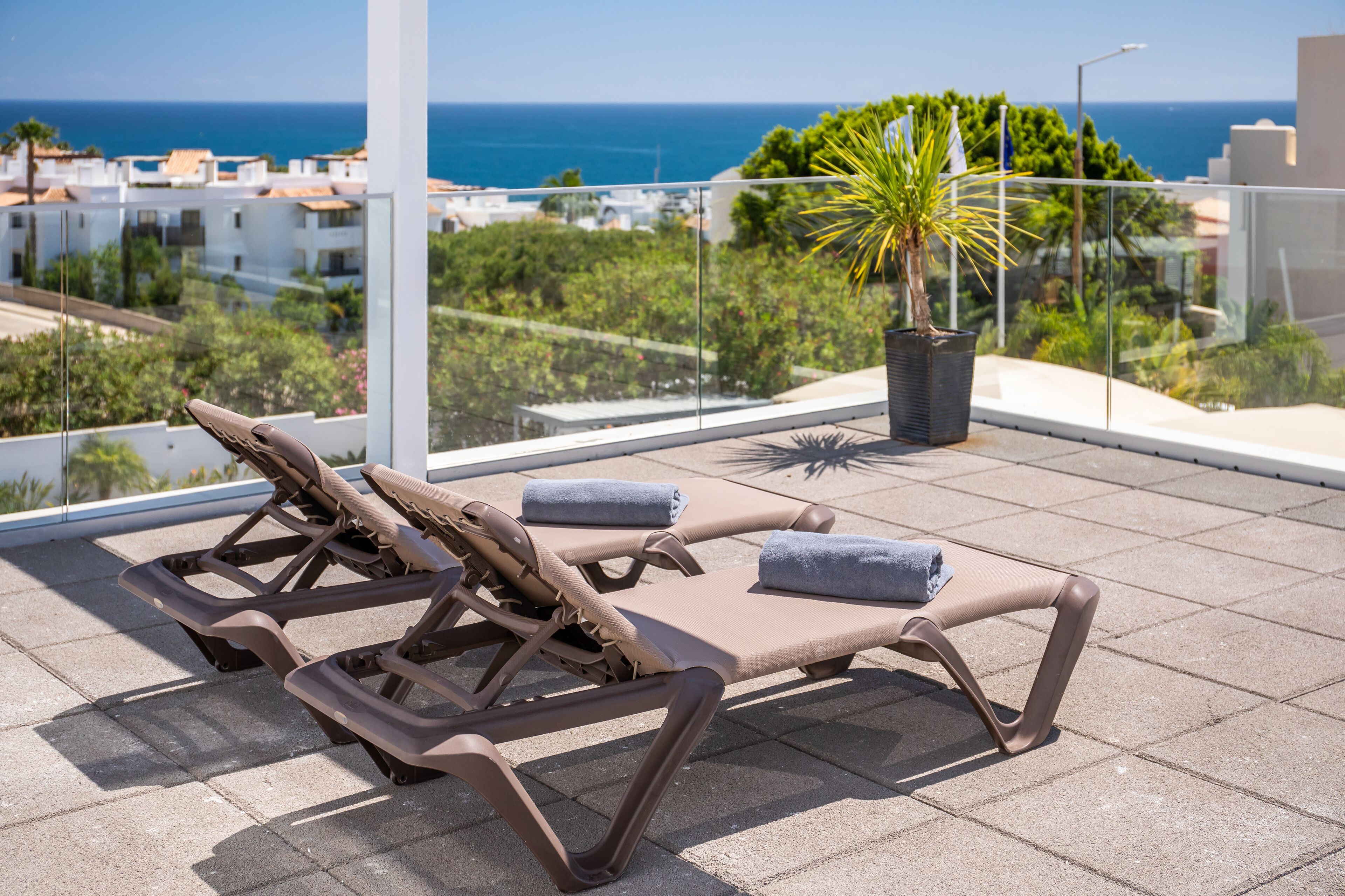 A rooftop terrace with two sun loungers, a potted plant, and a view of the sea and buildings.