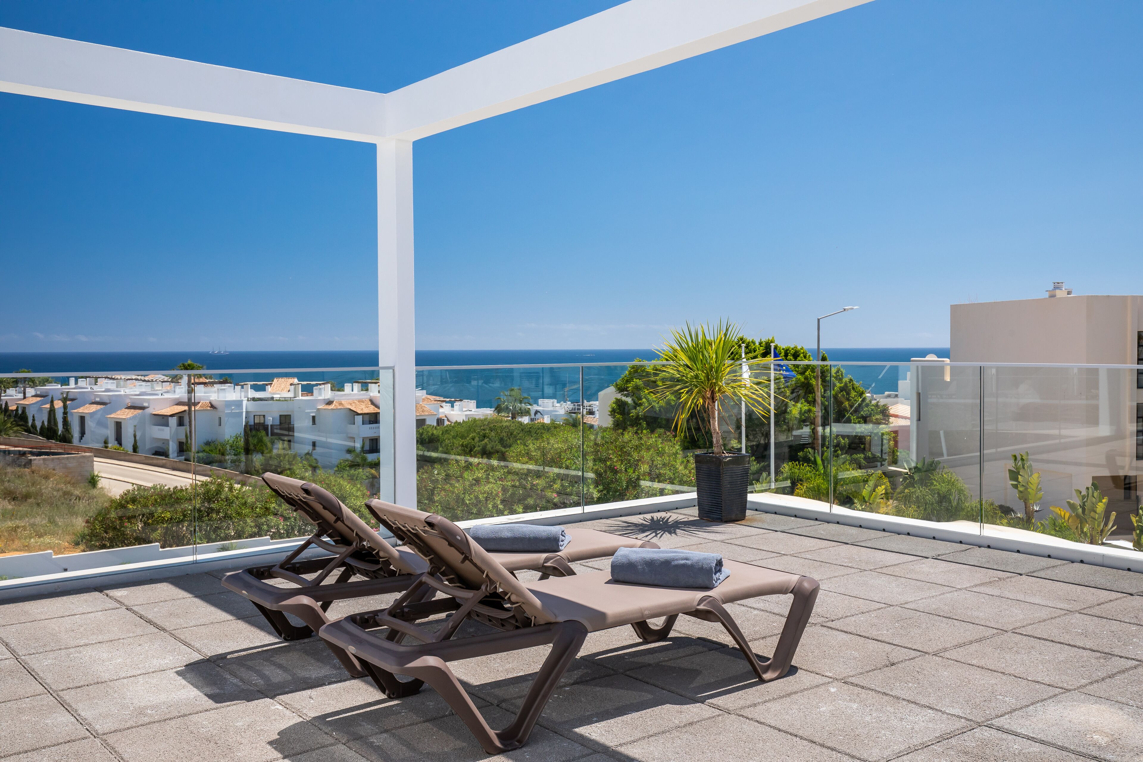 A rooftop terrace with sun loungers and a potted plant, overlooking a coastal cityscape.