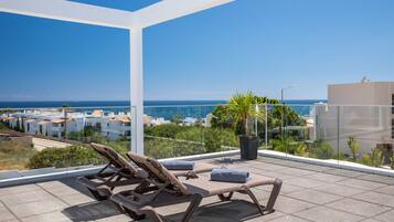 A rooftop terrace with sun loungers and a potted plant, overlooking a coastal cityscape.