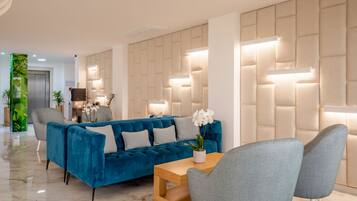 Un vestíbulo moderno con un sofá azul, sillones grises, una mesa de centro de madera y una pared con paneles iluminados.