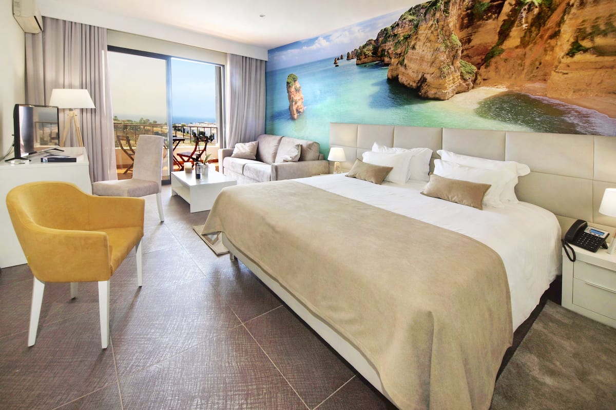 A hotel room with a large bed, a sofa, a chair, a desk, and a painting of a coastal scene.