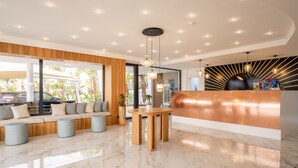 Un espace d’accueil moderne avec un comptoir de réception en bois, un coin salon avec un canapé et des poufs, et une grande fenêtre donnant sur l’extérieur.