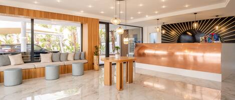Un espace d’accueil moderne avec un comptoir de réception en bois, un coin salon avec un canapé et des poufs, et une grande fenêtre donnant sur l’extérieur.