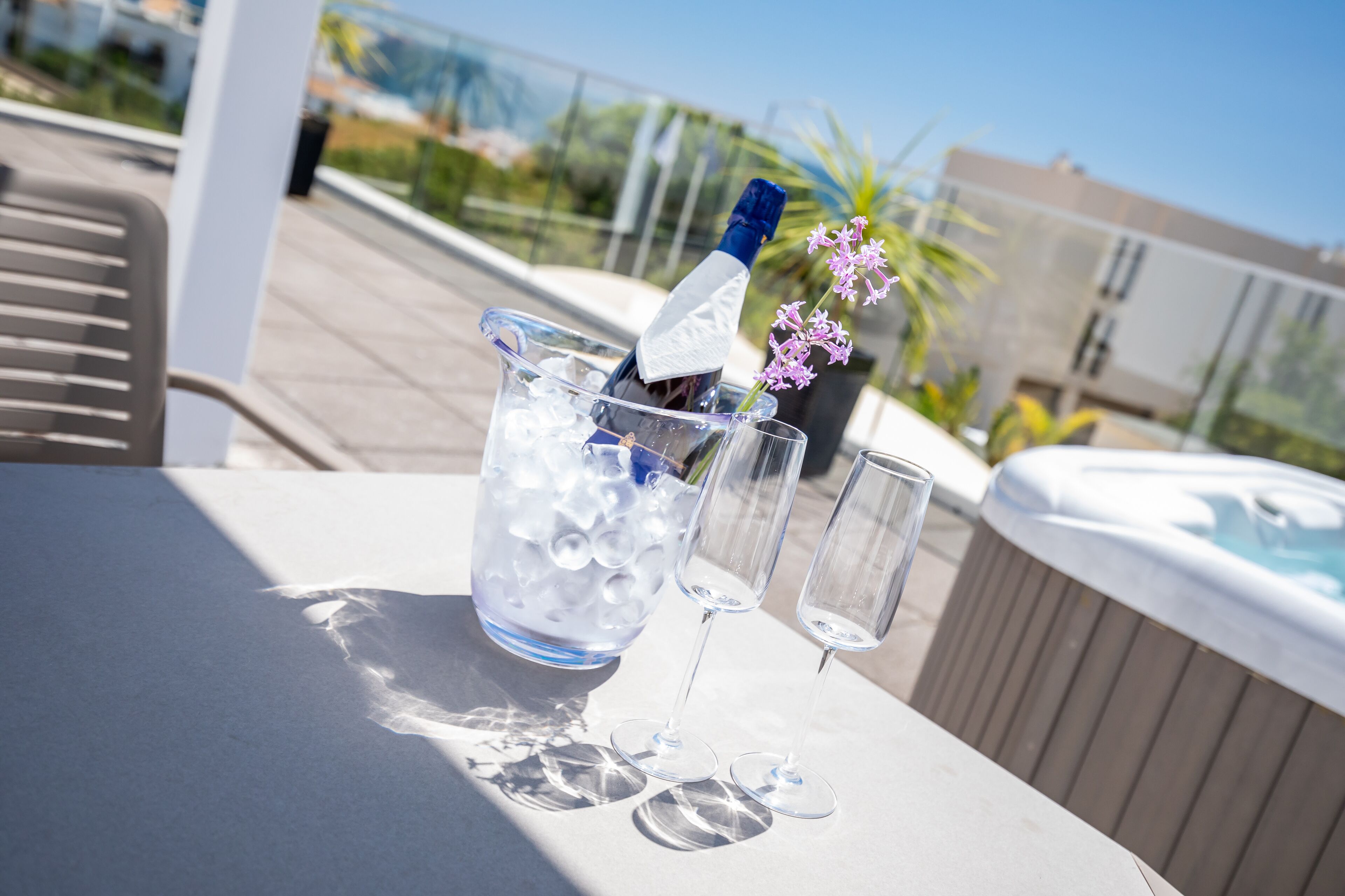A table with a champagne bottle, ice, and two glasses on a terrace with a hot tub and a view of buildings in the background.