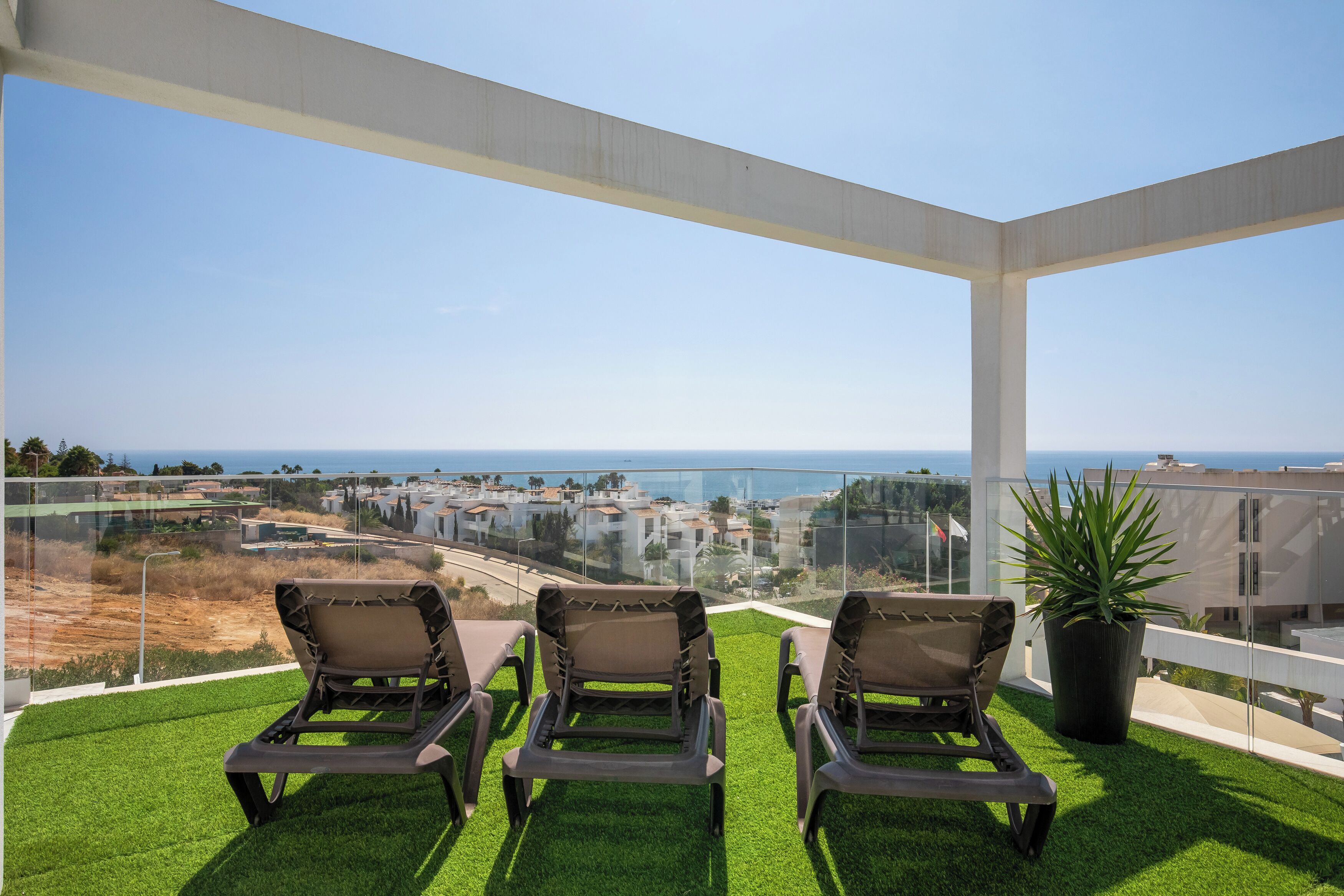 A balcony with three lounge chairs, a potted plant, and a view of the sea and buildings.