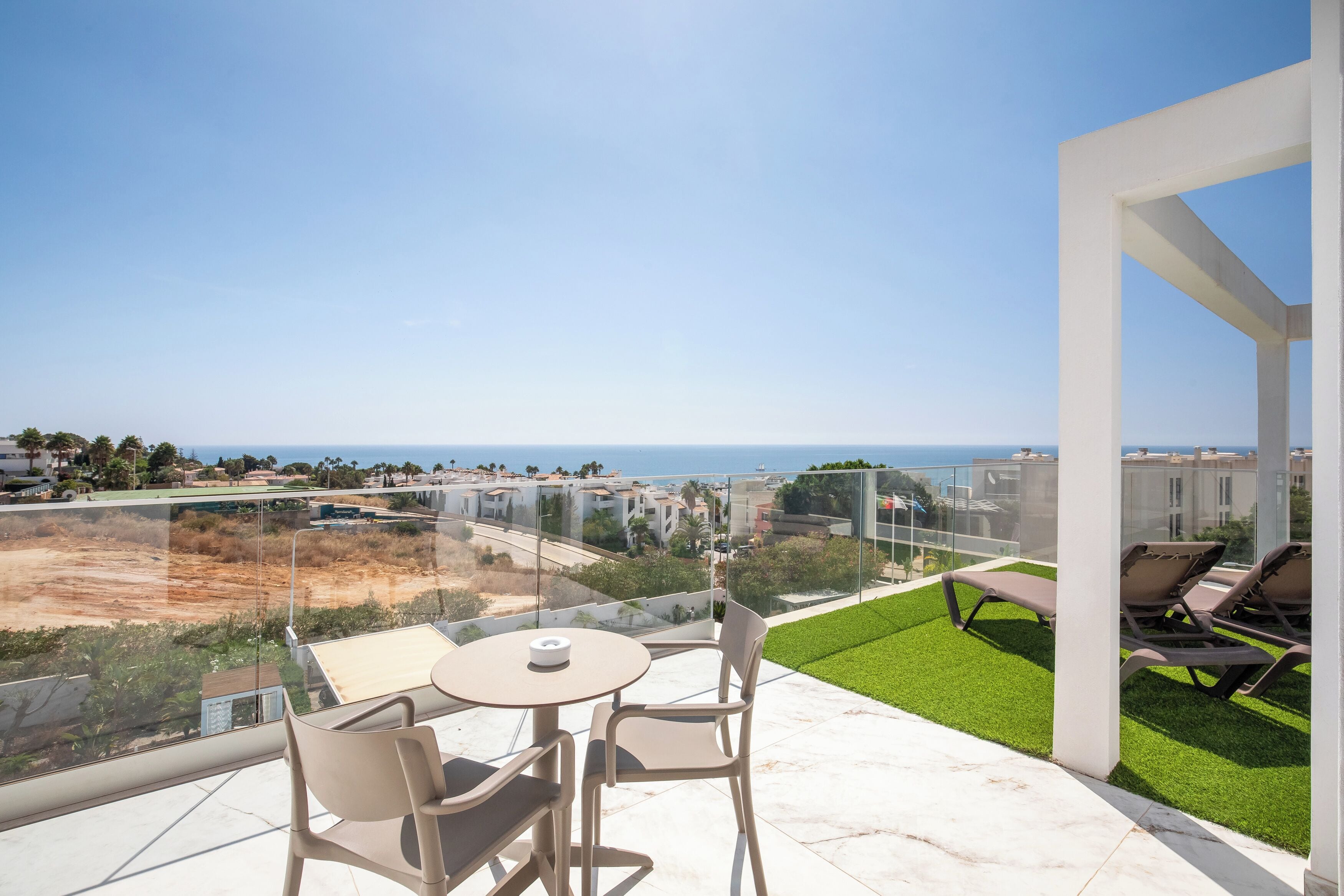 A balcony with a table and chairs, a grassy area, and a view of the sea and buildings.