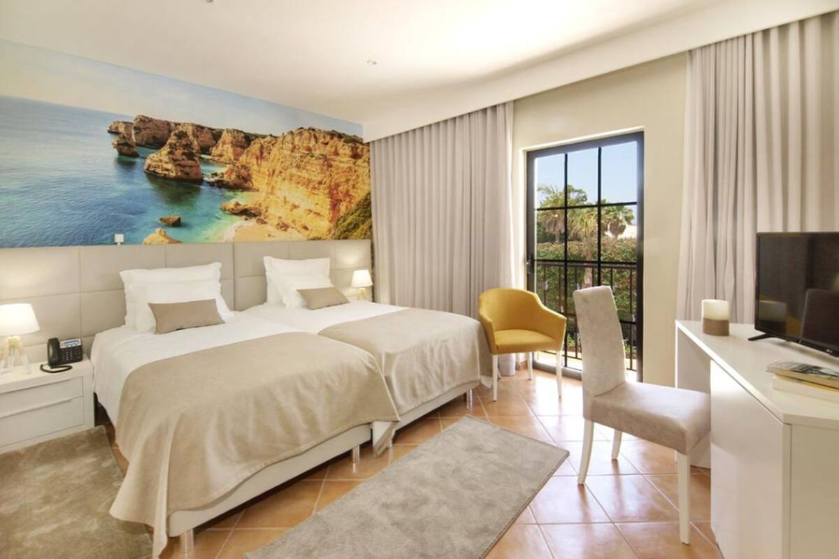 A hotel room with two beds, a desk with a TV, a chair, a balcony with curtains, and a painting of a coastal landscape.