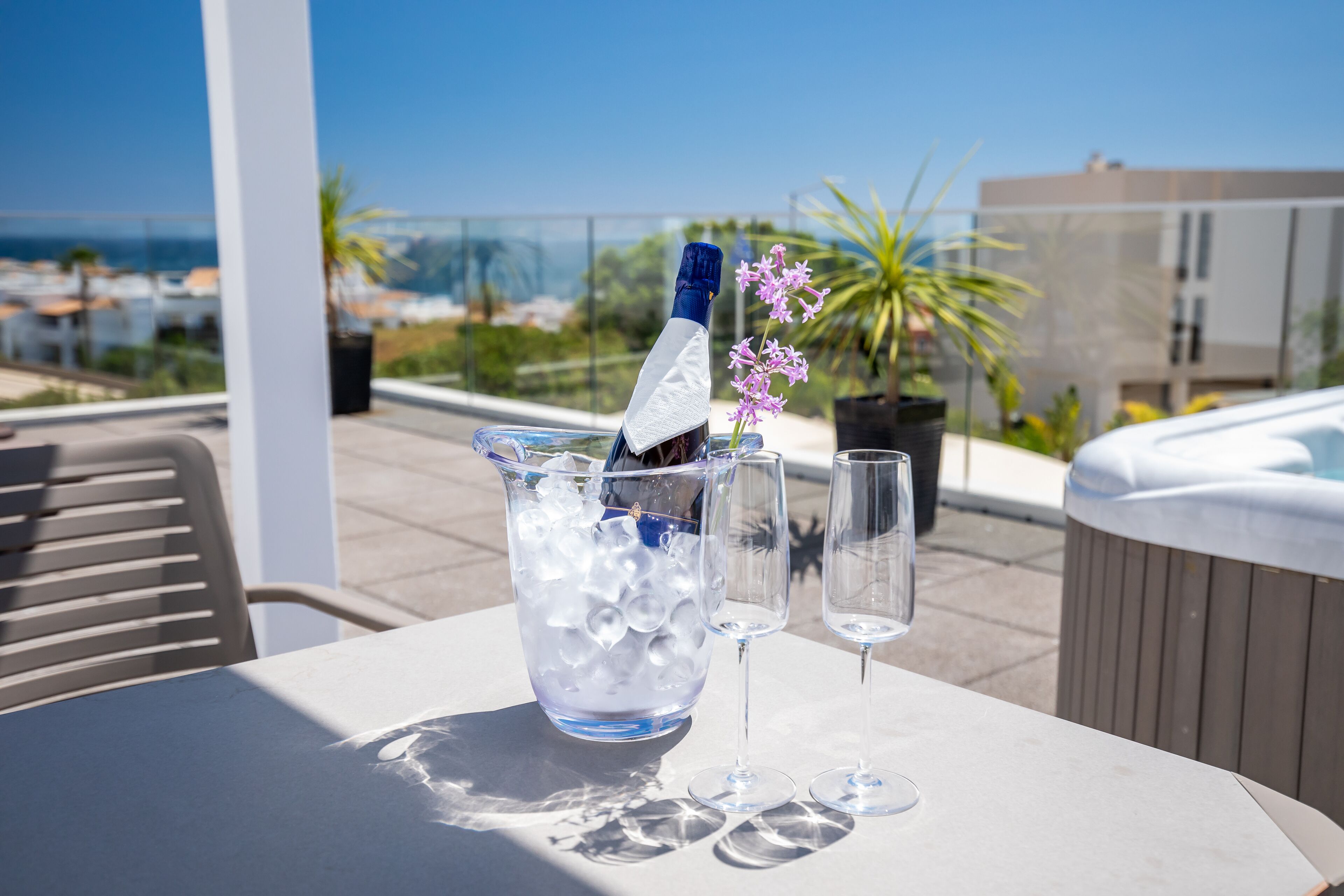 A table with a bottle of champagne, ice, and two glasses on a terrace with a view of the sea and buildings.