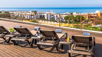 Un salón en la azotea con sillones de sol, una mesa con una bebida y vistas al océano y edificios.
